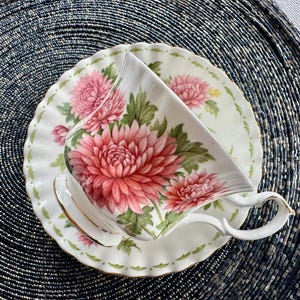 May include: White teacup and saucer set with a floral design. The teacup and saucer are decorated with large pink chrysanthemums and green leaves. The edges of the saucer and cup are scalloped and trimmed with gold.