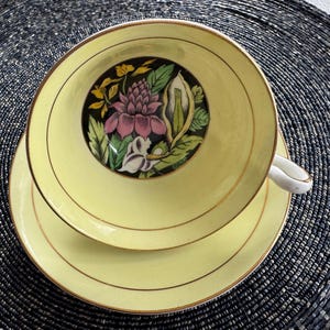 Puede incluir: Una taza de té y un platillo amarillos con un diseño floral. El diseño presenta una flor rosa con hojas verdes y una flor blanca con un centro amarillo.