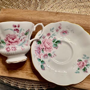May include: A white teacup and saucer set with a floral pattern featuring pink and green roses. The set has a gold trim around the edges.