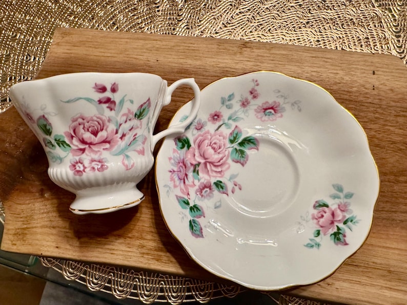May include: A white teacup and saucer set with a floral pattern featuring pink and green roses. The set has a gold trim around the edges.