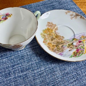 May include: A white porcelain teacup and saucer set featuring a delicate landscape illustration with a stone bridge, trees, and colorful flowers. The teacup has a light green handle and gold trim along the scalloped edges of both pieces.