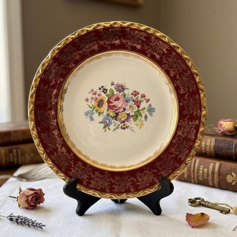 May include: A vintage decorative plate with a maroon border and gold filigree design. The center features a floral bouquet in shades of pink, yellow, blue, and purple. The plate is displayed on a black stand, showcasing its elegant design.