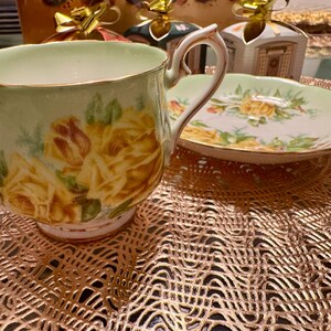 May include: A vintage teacup and saucer set with a delicate floral design. The cup and saucer are both white with a light green background and feature yellow roses with green leaves. The cup has a gold rim and a handle.