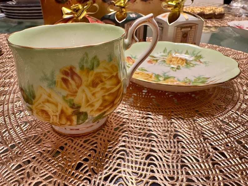 May include: A vintage teacup and saucer set with a delicate floral design. The cup and saucer are both white with a light green background and feature yellow roses with green leaves. The cup has a gold rim and a handle.