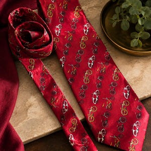 May include: A deep red necktie featuring a repeating pattern of small bicycles in yellow, black, and white. The tie has a smooth, slightly reflective finish and is displayed with the narrow end coiled and the wide end laid flat to show the print.