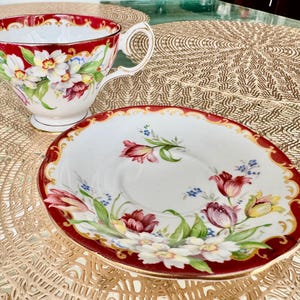 May include: A vintage teacup and saucer set. The teacup and saucer are white with a red border and floral design. The teacup has a handle and sits next to the saucer. The floral design includes white, red, and yellow flowers.