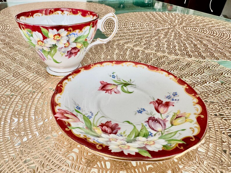 May include: A vintage teacup and saucer set. The teacup and saucer are white with a red border and floral design. The teacup has a handle and sits next to the saucer. The floral design includes white, red, and yellow flowers.