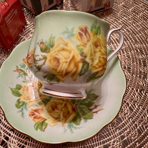 May include: A green and white teacup and saucer set with a floral design featuring yellow roses. The teacup has a delicate handle and the saucer has a scalloped edge.