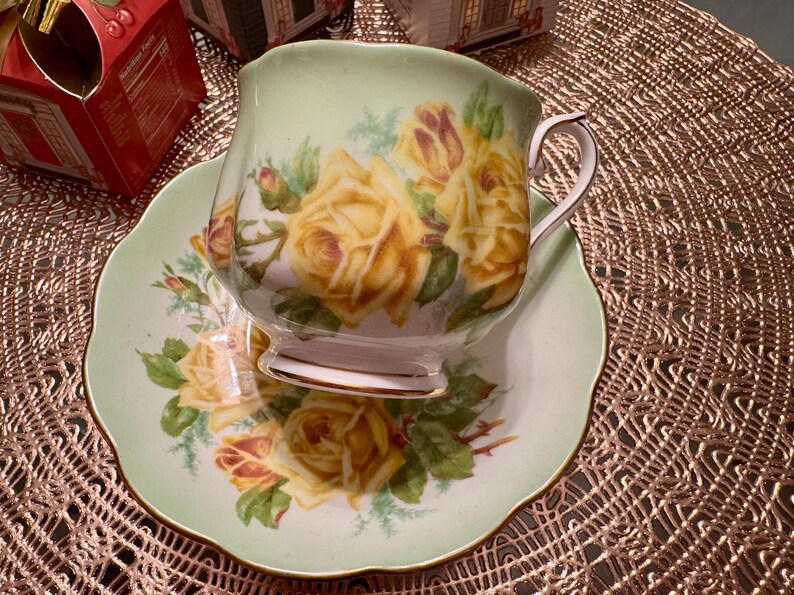 May include: A green and white teacup and saucer set with a floral design featuring yellow roses. The teacup has a delicate handle and the saucer has a scalloped edge.
