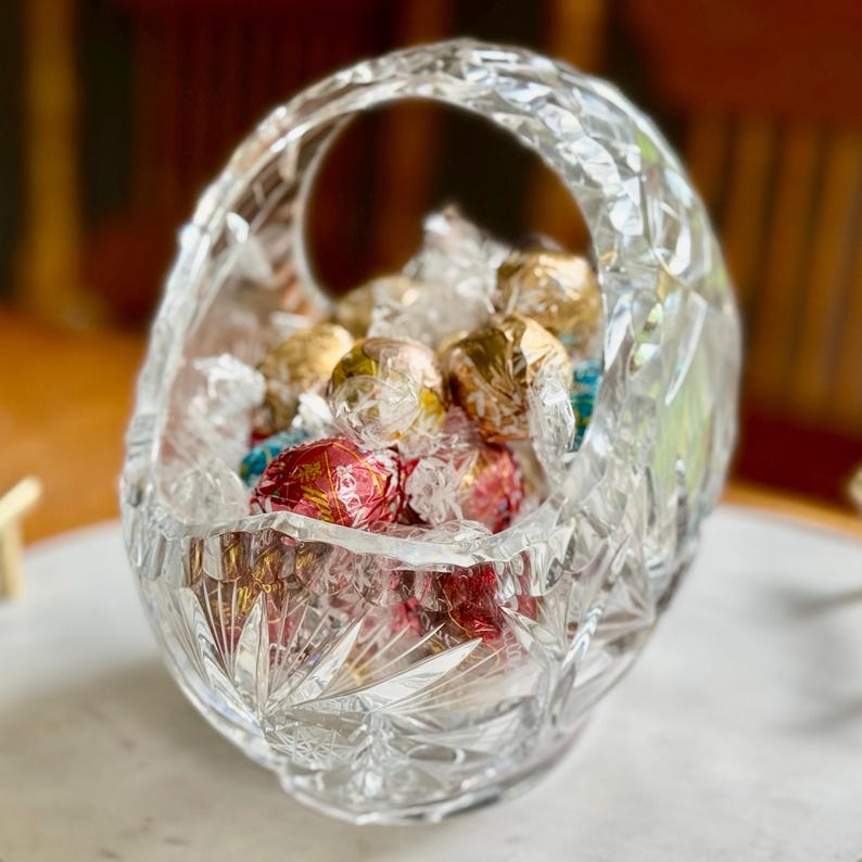 May include: A clear, cut-glass basket with a handle, filled with wrapped candies in gold, red, and blue foil. The basket has a starburst pattern on the base and a diamond-cut design on the sides.