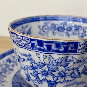 May include: Close-up of a white teacup and saucer with a blue floral pattern and gold trim. The teacup has a faceted design and a geometric border. The saucer is partially visible, also featuring the blue floral design.