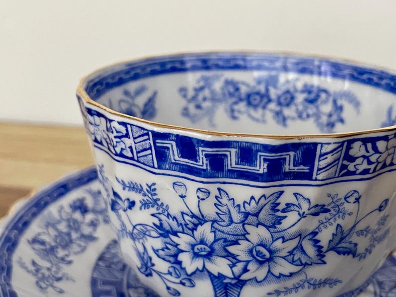 May include: Close-up of a white teacup and saucer with a blue floral pattern and gold trim. The teacup has a faceted design and a geometric border. The saucer is partially visible, also featuring the blue floral design.