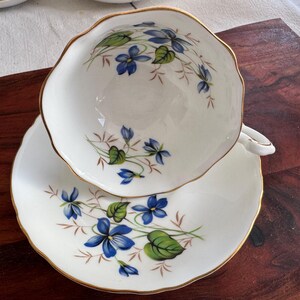 May include: A white teacup and saucer set with a delicate floral pattern featuring blue and green violets. The cup and saucer have a gold trim around the edges.