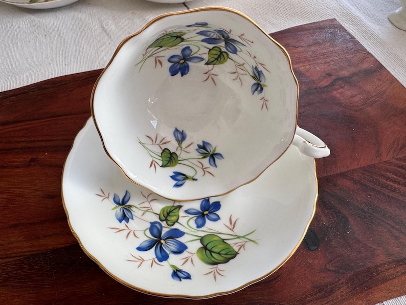 May include: A white teacup and saucer set with a delicate floral pattern featuring blue and green violets. The cup and saucer have a gold trim around the edges.