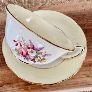 May include: A white teacup and saucer with a floral design. The cup has a gold rim and the saucer has a scalloped edge.