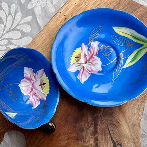 Puede incluir: Un juego de taza y platillo de t&eacute; azul y blanco con un dise&ntilde;o floral. La taza y el platillo presentan una orqu&iacute;dea rosa y amarilla con detalles dorados.