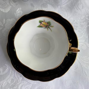 Pode incluir: Um conjunto de ch&aacute;vena e pires em porcelana branca com rebordo recortado e acabamento dourado. O interior da ch&aacute;vena apresenta um pequeno motivo de bot&atilde;o de rosa amarelo, enquanto o pires tem um acabamento em esmalte preto profundo.
