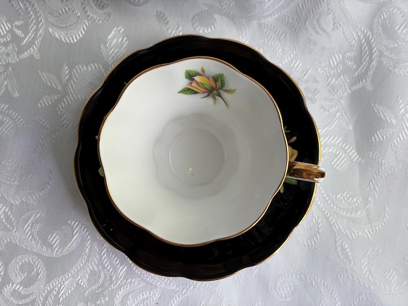 Pode incluir: Um conjunto de ch&aacute;vena e pires em porcelana branca com rebordo recortado e acabamento dourado. O interior da ch&aacute;vena apresenta um pequeno motivo de bot&atilde;o de rosa amarelo, enquanto o pires tem um acabamento em esmalte preto profundo.