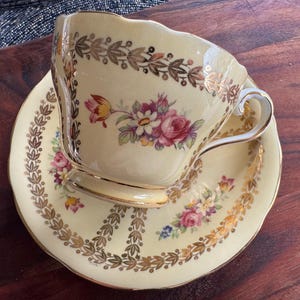 May include: A cream-colored porcelain teacup and saucer set featuring a delicate floral bouquet of pink, yellow, and purple flowers. The rim and base are decorated with a gold leaf laurel leaf pattern and gold trim on the handle and edges.
