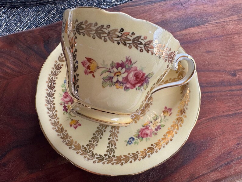 May include: A cream-colored porcelain teacup and saucer set featuring a delicate floral bouquet of pink, yellow, and purple flowers. The rim and base are decorated with a gold leaf laurel leaf pattern and gold trim on the handle and edges.