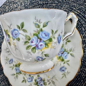 Pu&ograve; includere: Una tazza da t&egrave; e un piattino bianchi con un motivo floreale blu e verde. La tazza ha un bordo dorato e il piattino ha un bordo ondulato.