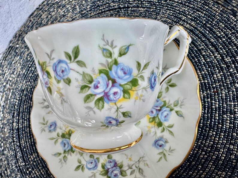 Pu&ograve; includere: Una tazza da t&egrave; e un piattino bianchi con un motivo floreale blu e verde. La tazza ha un bordo dorato e il piattino ha un bordo ondulato.