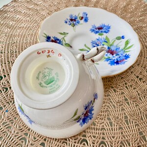 May include: A white teacup and saucer with a blue and white floral pattern. The teacup has a gold rim and the saucer has a gold band. The bottom of the teacup has a green and white label that reads "Bone China, Hammersley & Co., Made in England".