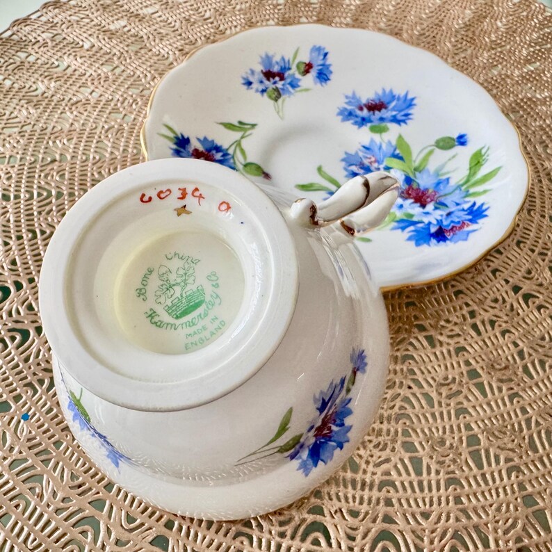 May include: A white teacup and saucer with a blue and white floral pattern. The teacup has a gold rim and the saucer has a gold band. The bottom of the teacup has a green and white label that reads "Bone China, Hammersley & Co., Made in England".