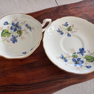 May include: A white teacup and saucer set with a blue and green floral pattern. The cup has a handle and the saucer has a scalloped edge.
