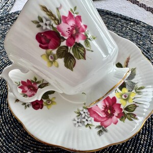 Puede incluir: Un juego de taza y platillo de t&eacute; blanco con un patr&oacute;n floral con flores rosas, rojas y amarillas. La taza y el platillo tienen un borde dorado.