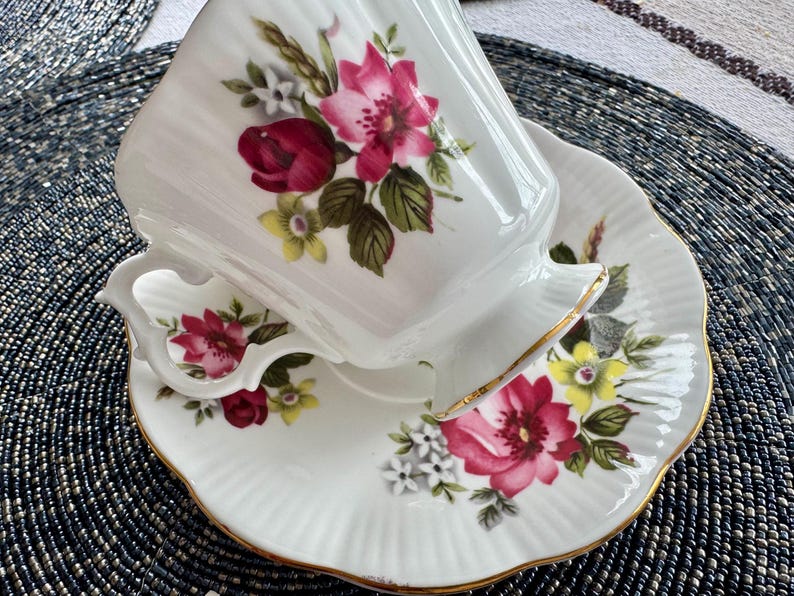 Puede incluir: Un juego de taza y platillo de t&eacute; blanco con un patr&oacute;n floral con flores rosas, rojas y amarillas. La taza y el platillo tienen un borde dorado.