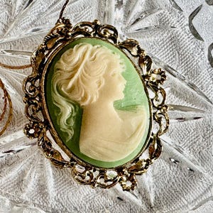 May include: Oval cameo pendant necklace with a gold-tone filigree frame. The cameo features a profile of a woman in ivory against a pale green background. The necklace has a delicate gold chain.