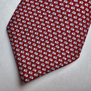 Puede incluir: Una corbata roja con un patr&oacute;n repetitivo de peque&ntilde;os elefantes blancos con orejas y detalles en azul claro. La tela tiene un tejido fino y texturizado, y la corbata tiene una forma cl&aacute;sica terminada en punta.