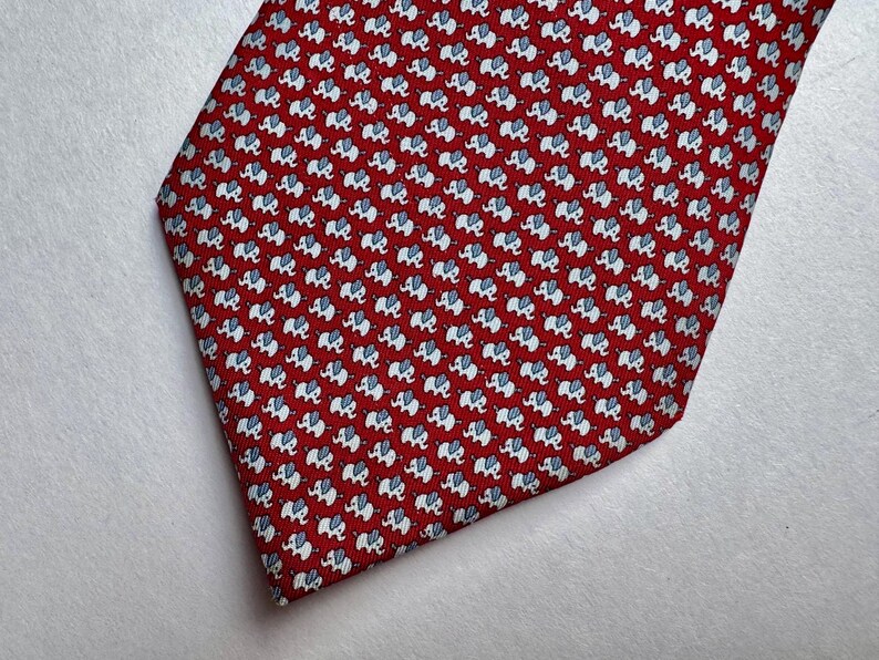 Puede incluir: Una corbata roja con un patr&oacute;n repetitivo de peque&ntilde;os elefantes blancos con orejas y detalles en azul claro. La tela tiene un tejido fino y texturizado, y la corbata tiene una forma cl&aacute;sica terminada en punta.