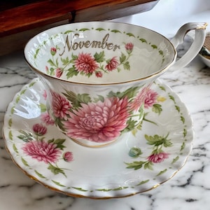 May include: A white teacup and saucer set with a floral design. The teacup and saucer feature pink chrysanthemums and green leaves. The word "November" is written in gold script on the teacup. The edges are trimmed with gold.