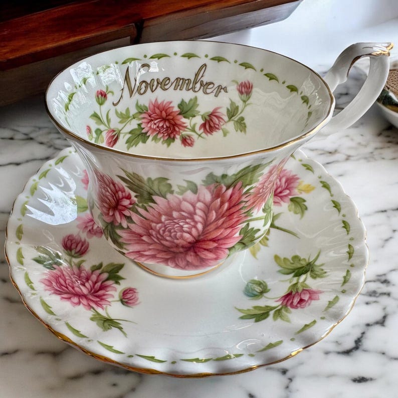 May include: A white teacup and saucer set with a floral design. The teacup and saucer feature pink chrysanthemums and green leaves. The word "November" is written in gold script on the teacup. The edges are trimmed with gold.