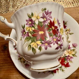 May include: A white teacup and saucer set with a floral design. The cup and saucer feature a delicate floral pattern with red, pink, yellow, and green flowers.