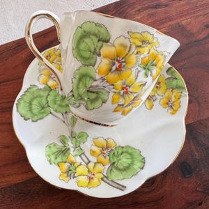 May include: A white teacup and saucer with a floral pattern. The pattern features yellow flowers with green leaves on a white background.