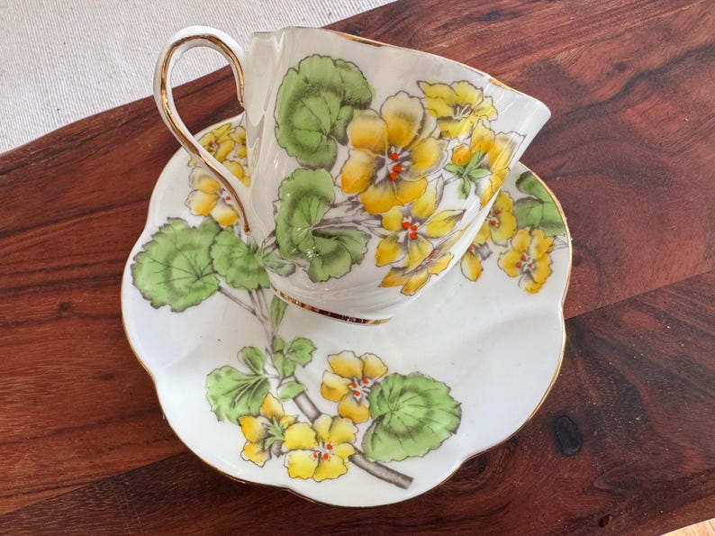 May include: A white teacup and saucer with a floral pattern. The pattern features yellow flowers with green leaves on a white background.