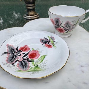 Puede incluir: Juego de taza y platillo de t&eacute; blanco con dise&ntilde;o floral. La taza y el platillo tienen un borde dorado. El dise&ntilde;o floral presenta flores rojas, rosas y negras con hojas verdes. El platillo tiene un borde festoneado.