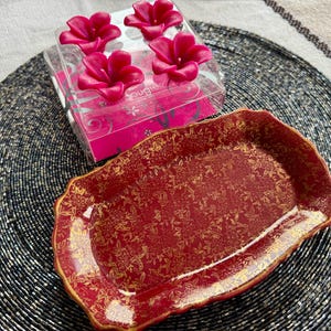 May include: A red and gold decorative serving tray with a floral pattern. The tray is sitting on a black woven placemat. There is a box of pink flower-shaped candles in the background.