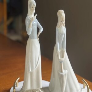 K&ouml;nnte beinhalten: Zwei Porzellanfiguren von Frauen in langen Kleidern. Eine h&auml;lt einen Vogel, die andere einen Regenschirm. Die Figuren sind in Wei&szlig;- und Hellblaut&ouml;nen gehalten und stehen auf einer marmor&auml;hnlichen Basis mit goldenen Griffen. Ein dekorativer Artikel.