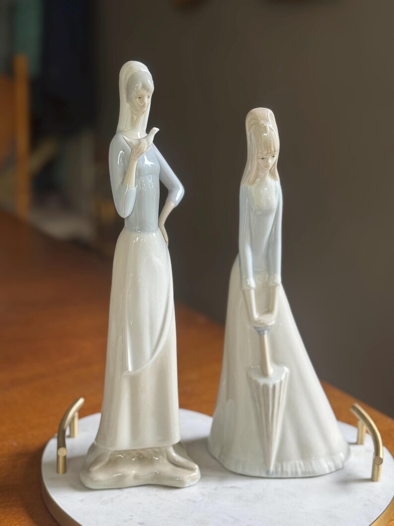K&ouml;nnte beinhalten: Zwei Porzellanfiguren von Frauen in langen Kleidern. Eine h&auml;lt einen Vogel, die andere einen Regenschirm. Die Figuren sind in Wei&szlig;- und Hellblaut&ouml;nen gehalten und stehen auf einer marmor&auml;hnlichen Basis mit goldenen Griffen. Ein dekorativer Artikel.