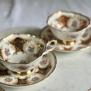May include: Two vintage teacups and saucers with gold trim and floral designs. The cups and saucers are white with gold accents and feature delicate pink and blue floral patterns. The set is displayed on a white tray with gold handles.