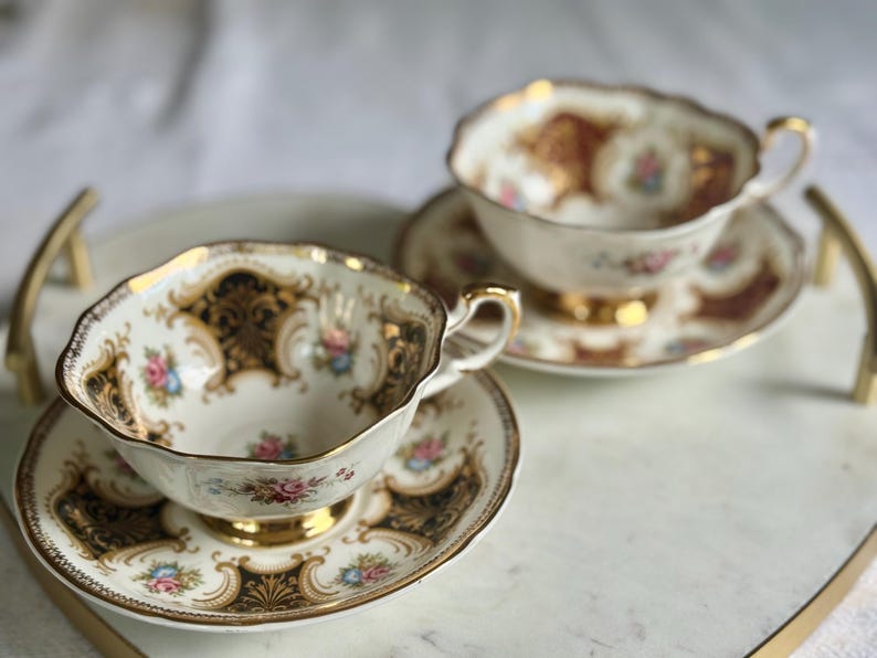May include: Two vintage teacups and saucers with gold trim and floral designs. The cups and saucers are white with gold accents and feature delicate pink and blue floral patterns. The set is displayed on a white tray with gold handles.