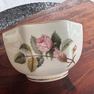 Pode incluir: Uma pequena tigela de cer&acirc;mica branca com uma borda dourada. A tigela &eacute; decorada com uma rosa rosa e folhas verdes. A tigela est&aacute; sobre uma superf&iacute;cie de madeira.