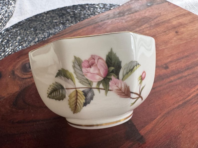 Pode incluir: Uma pequena tigela de cer&acirc;mica branca com uma borda dourada. A tigela &eacute; decorada com uma rosa rosa e folhas verdes. A tigela est&aacute; sobre uma superf&iacute;cie de madeira.