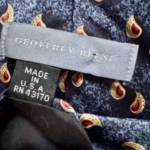 May include: A blue necktie featuring a repeating cream and red paisley pattern. A gray label reads GEOFFREY BEENE, and a black tag below it states MADE IN U.S.A. RN 43170. The fabric has a textured, mottled appearance throughout the design.