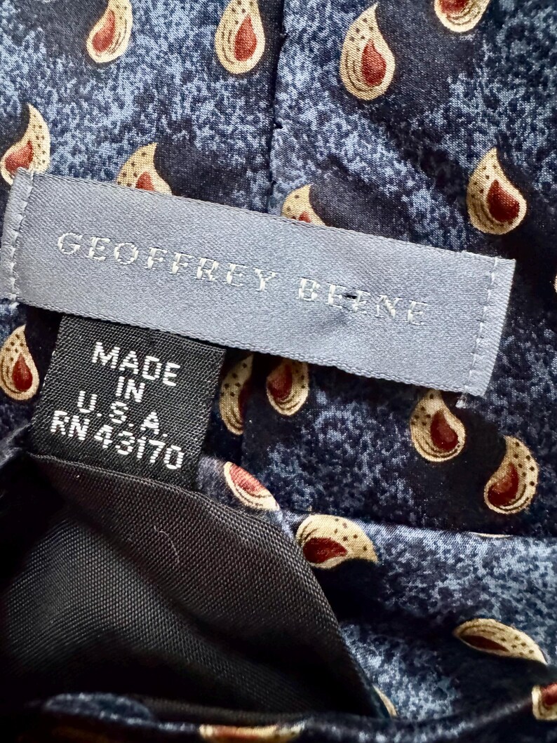 May include: A blue necktie featuring a repeating cream and red paisley pattern. A gray label reads GEOFFREY BEENE, and a black tag below it states MADE IN U.S.A. RN 43170. The fabric has a textured, mottled appearance throughout the design.
