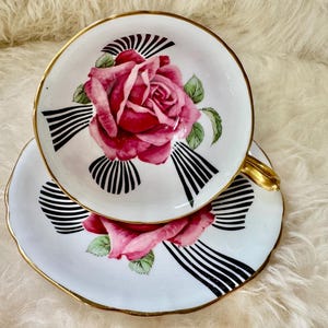 May include: A vintage teacup and saucer set. The white porcelain cup and saucer feature a large pink rose design with green leaves and black and white striped accents. Gold trim edges the cup and saucer.